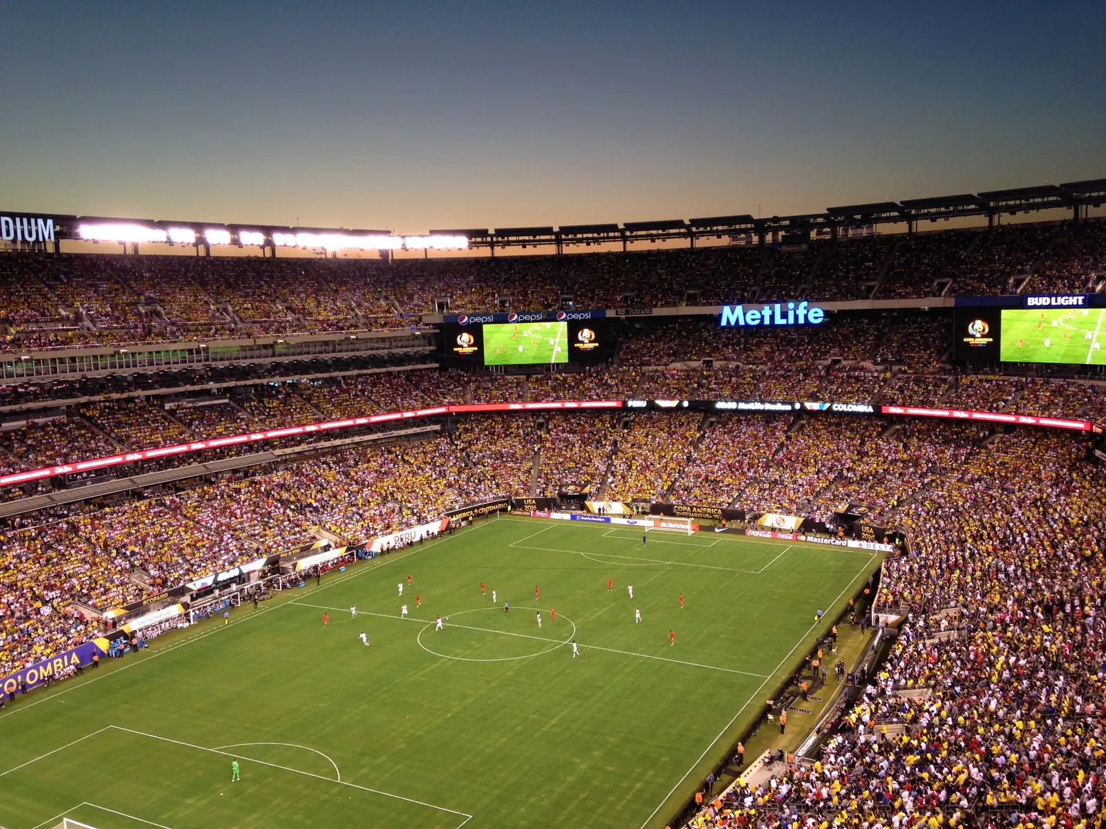 A crowd of football fans cheering in a stadium, with a large screen showing a football match.