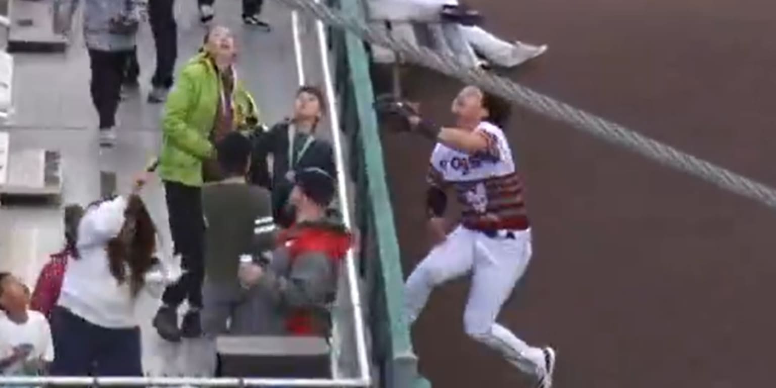 Baseball player Luis Suisbel leaps and extends his glove towards the top of an outfield wall.