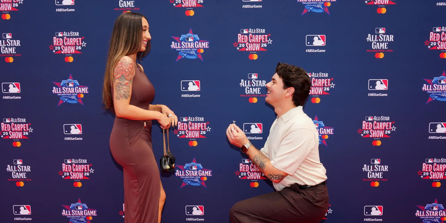 A couple embraces on the MLB All-Star red carpet, with the man kneeling in a proposal gesture.